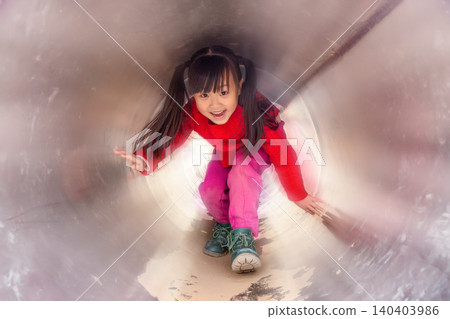a happy girl in a red sweater is crouching inside a metal tunnel in the playground. a happy girl in a red sweater is crouching inside a metal tunnel in the playground. 140403986
