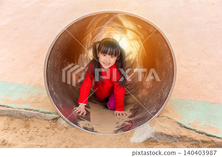 a happy girl in a red sweater is crouching inside a metal tunnel in the playground. a happy girl in a red sweater is crouching inside a metal tunnel in the playground. 140403987