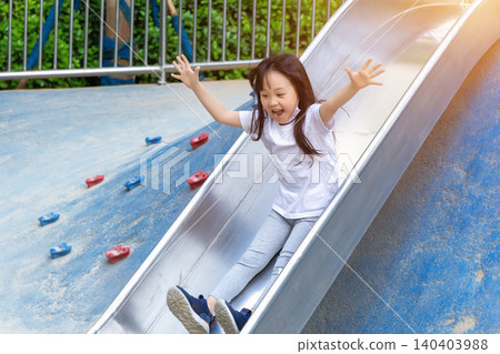 a cheerful girl sliding down a steel slide in the playground with open arms. a cheerful girl sliding down a steel slide in the playground with open arms. 140403988