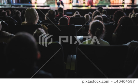 People sitting in rows, enjoying a show. Audience views the stage in a dim auditorium People sitting in rows, enjoying a show. Audience views the stage in a dim auditorium 140404597