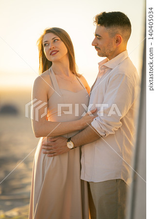 Couple stands together at sunset on the beach holding each other and smiling as the sun sets in the background near the ocean waves Couple stands together at sunset on the beach holding each other and smiling as the sun sets in the background near the ocean waves 140405034