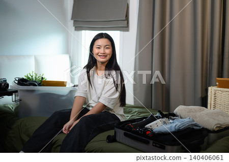 Happy Young Asian Woman Sitting on Bed with Open Suitcase Packing Belongings for University Dorm Room Happy Young Asian Woman Sitting on Bed with Open Suitcase Packing Belongings for University Dorm Room 140406061