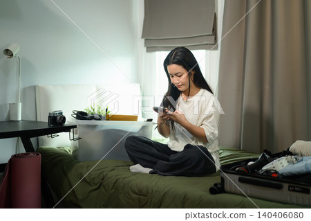 Happy Asian Student Sitting on Bed Using Smartphone While Packing Suitcase for University Happy Asian Student Sitting on Bed Using Smartphone While Packing Suitcase for University 140406080