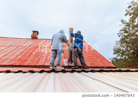 Two men worker installing metal chimney on red house roof. Professional roofers doing repair of smoke ventilation system. Maintenance service and external building construction work. Two men worker installing metal chimney on red house roof. Professional roofers doing repair of smoke ventilation system. Maintenance service and external building construction work. 140407843