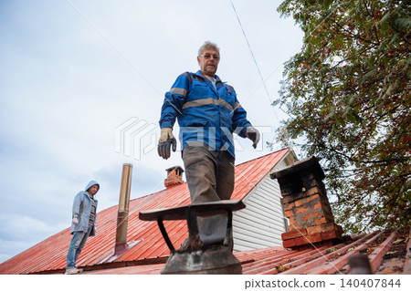 Chimney sweep working on house roof. Professional man repairing building ventilation system. Maintenance service for heater and fireplace. Construction worker fixing roof chimney exterior. Chimney sweep working on house roof. Professional man repairing building ventilation system. Maintenance service for heater and fireplace. Construction worker fixing roof chimney exterior. 140407844