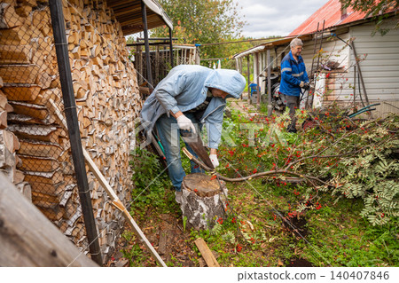 Two men working in courtyard to clear cut rowan branches. Seasonal garden maintenance and tree pruning. Helpers organizing wood and gathering garden waste in backyard for cleanup. Two men working in courtyard to clear cut rowan branches. Seasonal garden maintenance and tree pruning. Helpers organizing wood and gathering garden waste in backyard for cleanup. 140407846