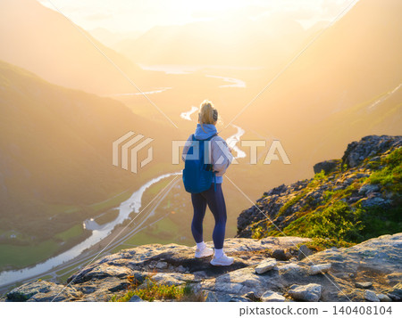 Athletic girl with a hiking backpack stands on a rock overlooking a sun-drenched mountain valley 140408104