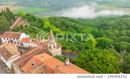 Famous medieval fortress town of Motovun stands proud on green hill in Istria. Aerial perspective shows ancient city walls, terracotta roofs, and mist rising from surrounding truffle forests. 140408332