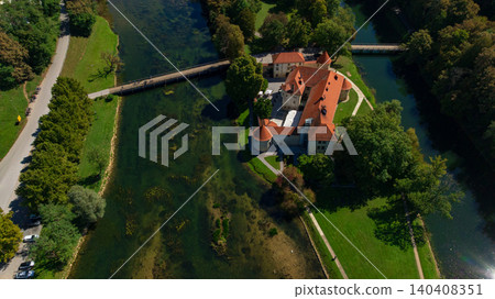 Stunning top down drone view captures medieval Otocec Castle situated on small island. Terracotta roofs and stone walls are surrounded by green trees and turquoise Krka river waters. Stunning top down drone view captures medieval Otocec Castle situated on small island. Terracotta roofs and stone walls are surrounded by green trees and turquoise Krka river waters. 140408351