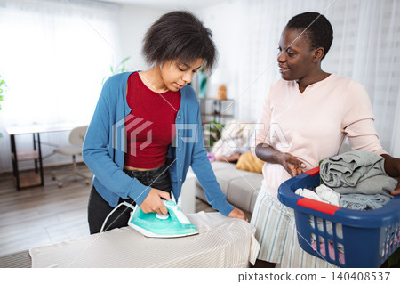 Mother watching daughter ironing clothes at home Mother watching daughter ironing clothes at home 140408537