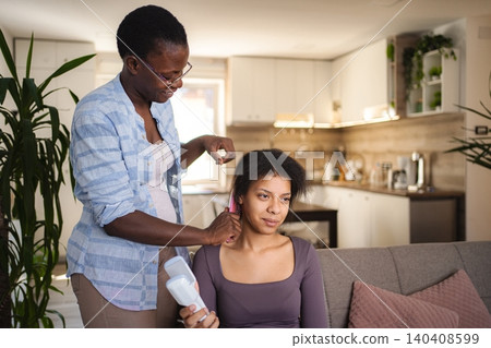 Mother combing her teenage daughter's hair on the sofa in the living room 140408599