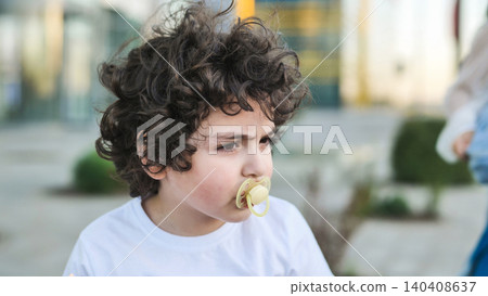 Young boy with curly hair thinking outdoors, having a pacifier in mouth Young boy with curly hair thinking outdoors, having a pacifier in mouth 140408637