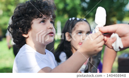 Young boy eagerly watching an adult unwrapping a popsicle for him outdoors Young boy eagerly watching an adult unwrapping a popsicle for him outdoors 140408948
