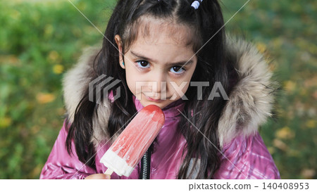 Little girl wearing hooded jacket enjoying frozen popsicle outdoors in park Little girl wearing hooded jacket enjoying frozen popsicle outdoors in park 140408953