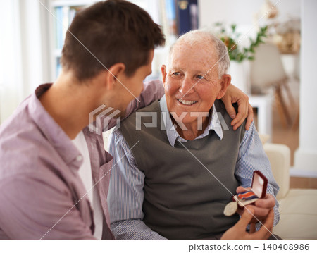 Father, adult son and medal for gift, support and congratulations with celebration and bonding at home. Men in living room, family time with reward or award for present, happiness and gratitude 140408986