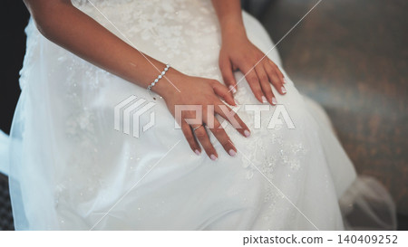Bride sitting on chair wearing elegant white wedding dress with floral embroidery, getting ready for wedding ceremony 140409252
