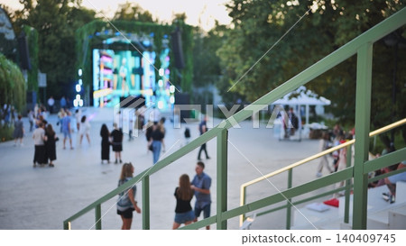 People gathering at an outdoor music festival with a bright stage in a park People gathering at an outdoor music festival with a bright stage in a park 140409745