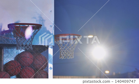 Basketball hoops with balls in a cage against a setting sun and bright stadium lights Basketball hoops with balls in a cage against a setting sun and bright stadium lights 140409747
