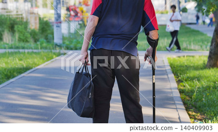 Elderly man walking with crutch and bag through a park in Moscow Elderly man walking with crutch and bag through a park in Moscow 140409903