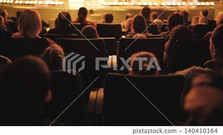 Spectators sitting in rows of chairs, observing a brightly lit stage during a show Spectators sitting in rows of chairs, observing a brightly lit stage during a show 140410164