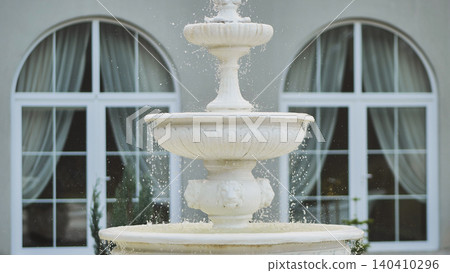 Cascading water flowing through multilevel white fountain, softening stone garden backdrop near blurred wedding venue arched windows Cascading water flowing through multilevel white fountain, softening stone garden backdrop near blurred wedding venue arched windows 140410296