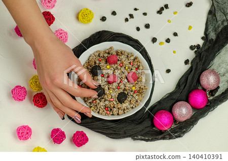 Contemporary kutia preparation with woman's hands, white bowl, blackberries, raspberries, colorful candy pearls, and festive decorations on white background. 140410391