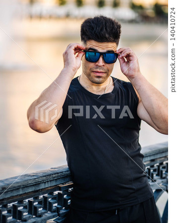 Confident man adjusting blue sunglasses outdoors against blurred serene background Confident man adjusting blue sunglasses outdoors against blurred serene background 140410724
