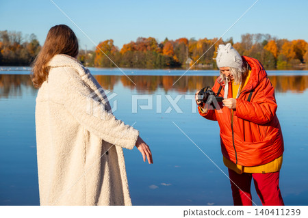 Woman photographer taking picture of another woman standing outdoors by lake. Creative female friend capturing portrait at autumn forest landscape. Leisure activity and hobby concept. 140411239