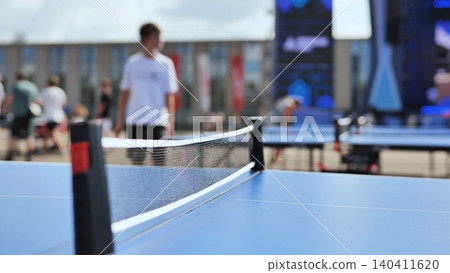 Blue table tennis table and net with blurred people playing ping pong outdoors Blue table tennis table and net with blurred people playing ping pong outdoors 140411620