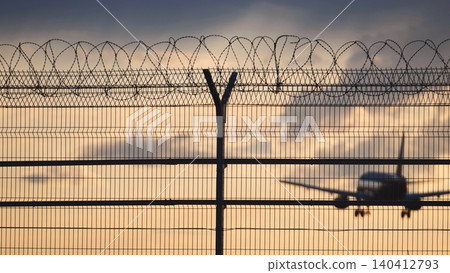 Airport fence with razor wire, an airplane landing in the background at sunset 140412793