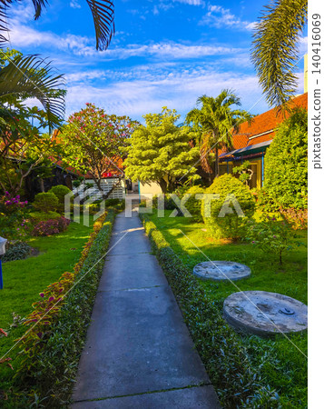 Beautiful tropical garden park featuring a paved walking path, green manicured lawns, and a blooming red flamboyant tree under a clear sunny blue sky. Beautiful tropical garden park featuring a paved walking path, green manicured lawns, and a blooming red flamboyant tree under a clear sunny blue sky. 140416069