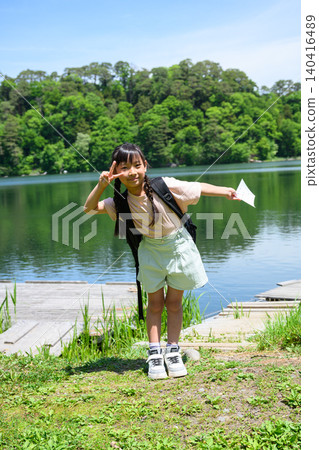 Elementary school girls on a field trip, with backpacks on their backs, smile and make peace signs by the lush green lakeside. 140416489