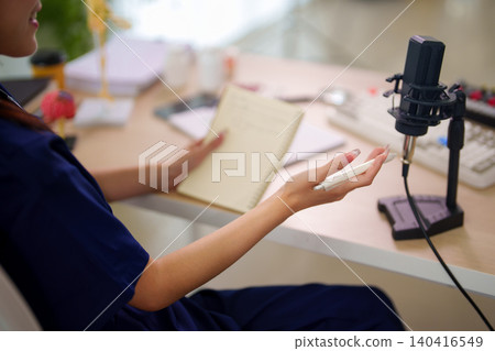Young Asian woman podcaster gesturing with hand while speaking into professional studio microphone during audio recording session at bright home workspace desk Young Asian woman podcaster gesturing with hand while speaking into professional studio microphone during audio recording session at bright home workspace desk 140416549