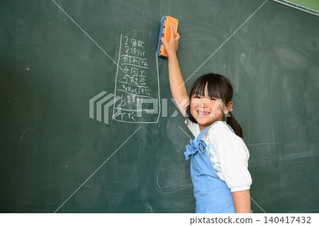A young elementary school girl smiles as she erases the blackboard in the classroom. 140417432