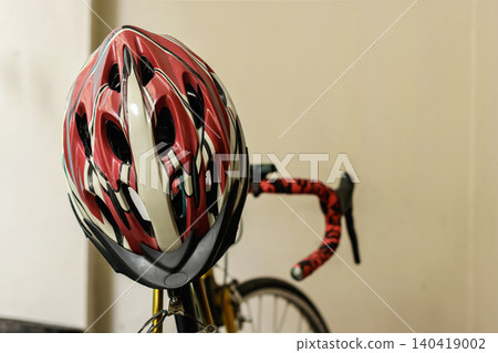 A close-up shot of a red, white, and black aerodynamic cycling safety helmet resting on the handlebars of a road bike indoors against a plain wall. High Quality Photo 140419002