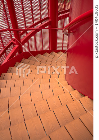 Looking down a vibrant red industrial spiral staircase featuring wooden steps and a mesh safety screen Looking down a vibrant red industrial spiral staircase featuring wooden steps and a mesh safety screen 140419035