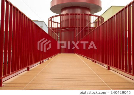 Low angle perspective of an outdoor walkway featuring wooden plank flooring and prominent red metal handrails Low angle perspective of an outdoor walkway featuring wooden plank flooring and prominent red metal handrails 140419045
