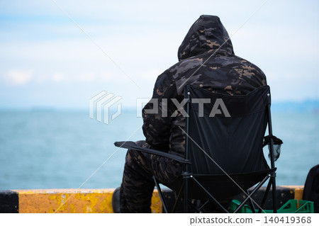 Rear view of a person wearing a camo hoodie sitting on a folding chair at a harbor pier Rear view of a person wearing a camo hoodie sitting on a folding chair at a harbor pier 140419368