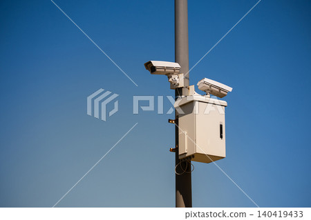 Two white CCTV surveillance cameras mounted on a gray pole against a clear blue sky background 140419433
