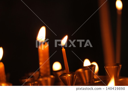 Close-up of glowing thin wax candles flickering against a dark, moody background in a temple Close-up of glowing thin wax candles flickering against a dark, moody background in a temple 140419898