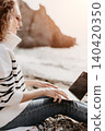 Laptop beach remote woman in striped sweater typing on a computer at a rocky coastline with mountains in background 140420350