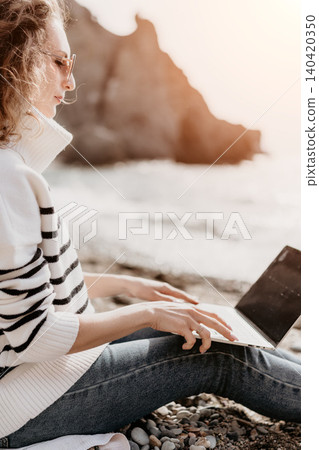 Laptop beach remote woman in striped sweater typing on a computer at a rocky coastline with mountains in background Laptop beach remote woman in striped sweater typing on a computer at a rocky coastline with mountains in background 140420350