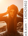 Woman Beanie Sunset, stylish female adjusts her knit cap while sitting on a rocky beach during the warm golden hour. 140420402