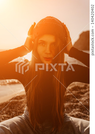 Woman Beanie Sunset, stylish female adjusts her knit cap while sitting on a rocky beach during the warm golden hour. Woman Beanie Sunset, stylish female adjusts her knit cap while sitting on a rocky beach during the warm golden hour. 140420402