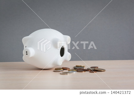 Coins scattered next to a white piggy bank on a wooden table in a simple setting Coins scattered next to a white piggy bank on a wooden table in a simple setting 140421372