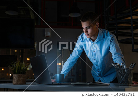 Young man works late at night in a modern office setting with a laptop and coffee Young man works late at night in a modern office setting with a laptop and coffee 140422781