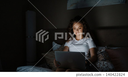 Woman sits on bed using laptop in dim light at home during evening hours Woman sits on bed using laptop in dim light at home during evening hours 140422824