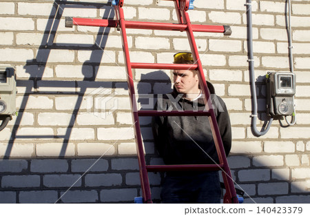 Worker standing by red ladder next to white brick wall. Electrician or technician man in safety glasses near electricity meter box, house renovation and construction site concept Worker standing by red ladder next to white brick wall. Electrician or technician man in safety glasses near electricity meter box, house renovation and construction site concept 140423379
