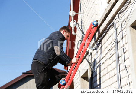 Worker climbing ladder on a house wall for installation or repair. Side view of young technician or electrician fixing cables outside building, house maintenance and telecommunication Worker climbing ladder on a house wall for installation or repair. Side view of young technician or electrician fixing cables outside building, house maintenance and telecommunication 140423382