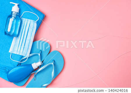 Coronavirus summer accessories. Blue towel, flip-flops, sunscreen, protective mask and hand sanitizer on pink Coronavirus summer accessories. Blue towel, flip-flops, sunscreen, protective mask and hand sanitizer on pink 140424016